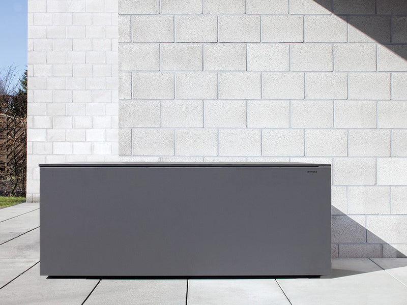 Graue Kissentruhe EL PECHO aus HPL-Material steht auf einer Terrasse vor einer hellen Ziegelwand. Die Truhe hat klare, minimalistische Linien und eine glatte Oberfläche. Sie hat keine sichtbaren Griffe oder Verzierungen, strahlt moderne Eleganz aus und bietet einen großzügigen Stauraum. Der Hintergrund zeigt eine ruhige Außenumgebung.