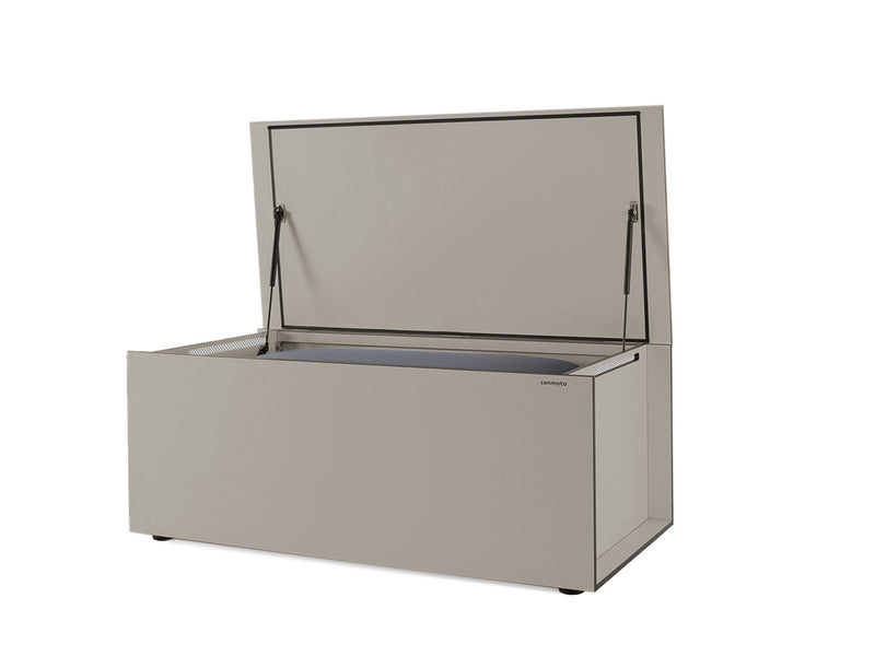 Beigefarbene Kissentruhe aus HPL-Material, rechteckig mit klaren Linien. Der geöffnete Deckel zeigt das Innenfach, unterstützt durch schwarze Gasdruckfedern. Links und rechts an der Truhe befinden sich Belüftungsschlitze. Die Truhe steht auf kleinen, runden Stellfüßen. Markenname "conmoto" ist rechts auf der Vorderseite zu sehen.