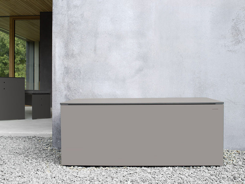 Graue, rechteckige Kissentruhe aus wetterbeständigem Material steht auf einem Kiesboden vor einer grauen Hauswand. Die glatte Oberfläche und klaren Linien betonen das minimalistische Design. Im Hintergrund ist ein offenes Fenster mit Holzrahmen zu erkennen.