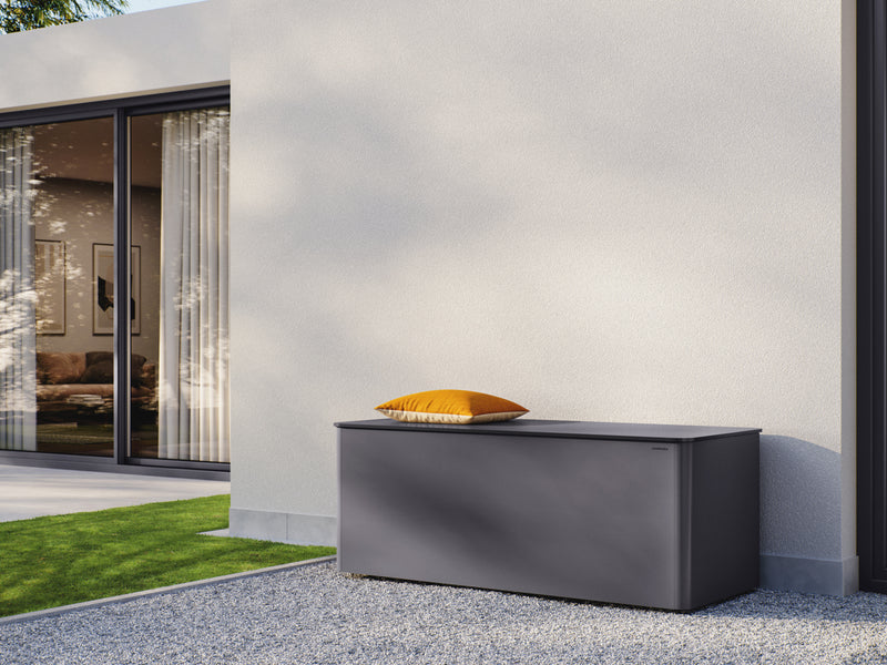 Moderne anthrazitfarbene RONDA Kissentruhe im Außenbereich vor einer hellen Hauswand platziert. Auf der Truhe liegt ein orangefarbenes Kissen. Die Oberfläche der Kissentruhe ist glatt, mit abgerundeten Kanten. Im Hintergrund befindet sich eine verglaste Terrassentür mit Blick ins Wohnzimmer. Der Boden um die Truhe besteht aus kleinen grauen Kieselsteinen, daneben ein kleiner Rasenstreifen.