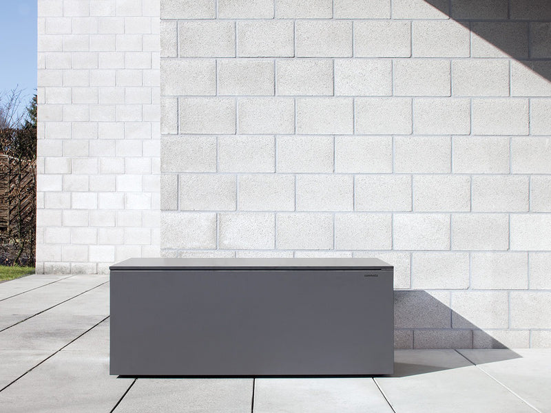 Graue Kissentruhe aus HPL-Material in rechteckiger Form, steht vor einer hellgrauen Steinwand. Die Oberfläche ist glatt und witterungsbeständig. Links neben der Truhe sind einige Pflanzen sichtbar, der Boden besteht aus großen, quadratischen Fliesen. Die Umgebung ist minimalistisch gestaltet und deutet ein modernes Außenbereichsdesign an.