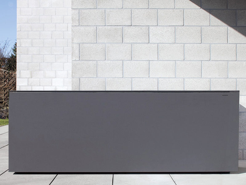 Graue Kissentruhe aus HPL-Material mit klaren Linien und minimalistischer Gestaltung steht vor einer hellen Steinmauer. Die Oberfläche ist glatt und witterungsbeständig, optimal für den ganzjährigen Außeneinsatz geeignet. Die Truhe besitzt eine schlichte, rechteckige Form und verspricht sowohl Funktionalität als auch modernes Design. Geradlinige Schatten deuten auf Sonneneinstrahlung von oben rechts hin.