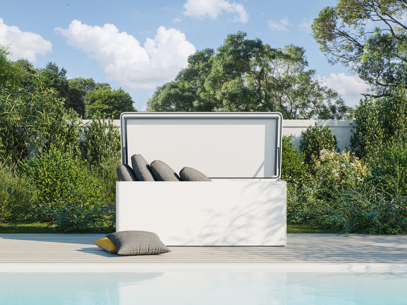 Weiße RONDA Kissentruhe mit geöffnetem Deckel auf einer Terrasse am Pool. Der Korpus aus wetterfestem Material hat abgerundete Ecken. Innen liegen mehrere graue Kissen ordnungsgemäß verstaut. Eine grüne Gartenlandschaft mit Büschen und Bäumen im Hintergrund ergänzt das Bild. Das Design der Truhe ist modern und schlicht, was gut zur Umgebung passt.
